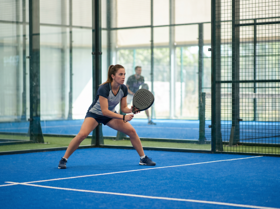 Padel player in a low ready stance preparing to return serve on a glass-walled court