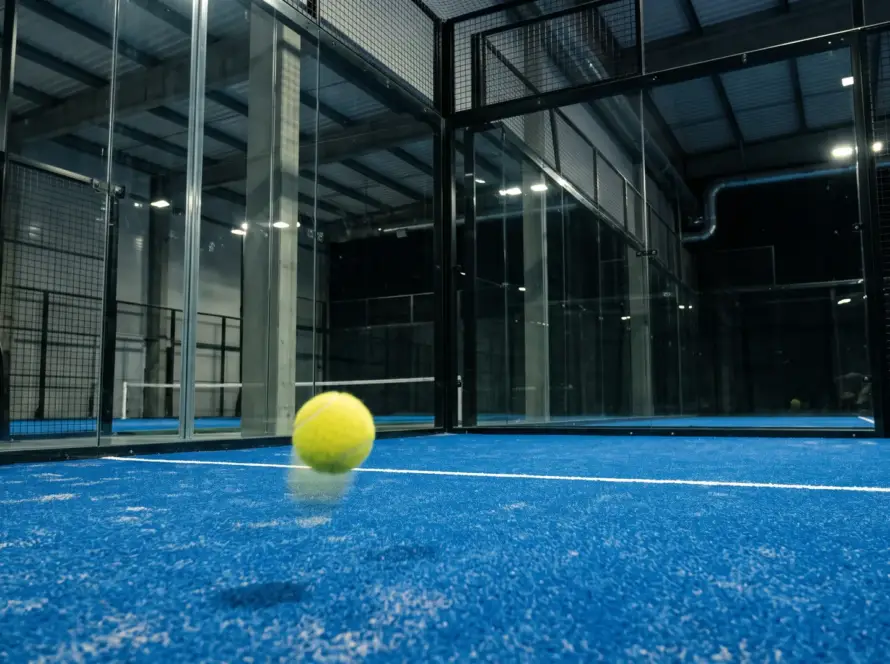 Yellow padel ball bouncing in front of the glass back wall of an indoor padel court