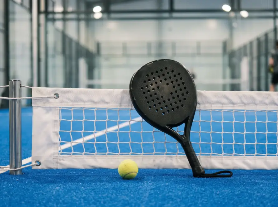 Padel racket resting against the net with a yellow ball on an indoor court